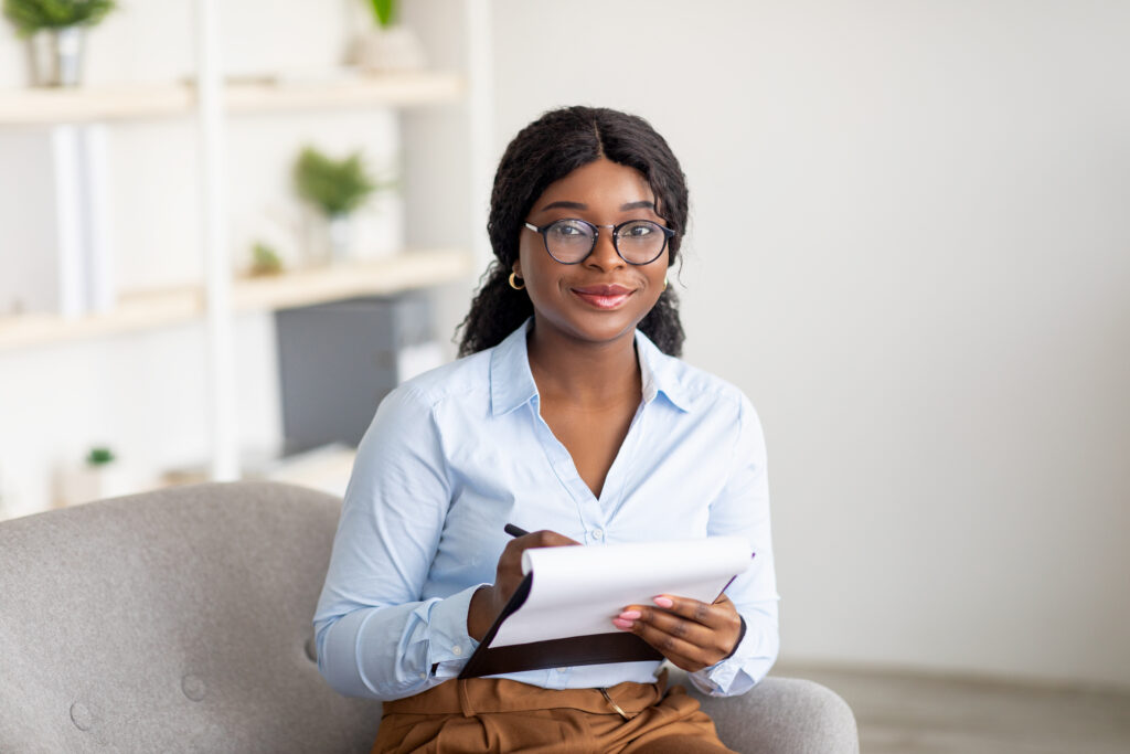 Friendly black female psychologist writing in clipboard, having session with client, sitting on couch at modern office. Happy psychotherapist taking notes during consultation in mental health clinic